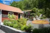 Trädgård med blommor, stenar, grusgångar och röda byggnader omgiven av grönska.