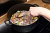 En hand rör om i en gjutjärnsgryta med kött och grönsaker på en spishäll.