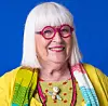 En leende kvinna med vitt hår, rosa glasögon och gul topp mot blå bakgrund.
