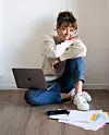 Kvinna sitter på golvet bredvid en laptop, papper och en mobil mot en ljus vägg.
