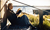 En kvinna sitter på en träterrass vid havet och läser en bok i solen.