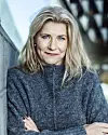 Blond kvinna i grå tröja med korsade armar framför en modern, oskarp bakgrund.