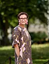 Kvinna med glasögon och rosa örhängen står utomhus framför en grön bakgrund.
