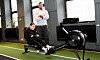 Två personer i ett gym, en sitter på en roddmaskin medan en står bredvid och tittar på.