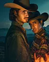 Två män i cowboyhatt står tätt intill varandra framför fjäll, med eld i förgrunden och blå kvällshimlen bakom.