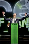 En man i kostym står bakom en grön talarstol och håller armarna upp i en scenmiljö.