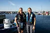 Två personer i flytvästar står i en blå gummibåt i en småbåtshamn med segelbåtar i bakgrunden.