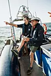 Två personer står på en båt och pekar ut över havet under en klar dag.