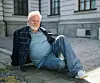 Äldre man sitter på marken utomhus framför en ljus byggnad.