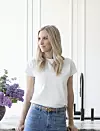 Kvinna i vit T-shirt och jeans står vid ett fönster i ett ljust rum.