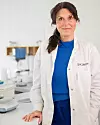 Person i vit labbrock och blå tröja står i ett laboratorium med laboratorieutrustning i bakgrunden.
