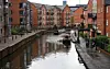 En smal båt färdas på en kanal mellan röda tegelbyggnader och gångvägar längs vattnet.