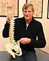 En man i svart tröja håller en modell av ryggrad och bäcken i ett rum med anatomiska planscher.