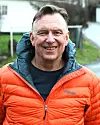Leende man i orange jacka står utomhus med suddig grön och grå bakgrund.