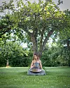 Kvinna sitter på gräset och spelar handpan under ett stort träd i en grön park.