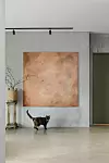Katt framför stort abstrakt konstverk i ett minimalistiskt rum med vas på stativ.