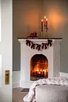Vit öppen spis med tända ljus och mörkröda dekorationer i ett vardagsrum.