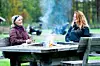 Två kvinnor sitter vid ett träbord utomhus med en liten eld i mitten och en mugg i handen.