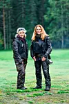 Två kvinnor i svarta motorcykelkläder står på en gräsmatta framför en mörk granskog.