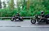 Två motorcyklister kör på en landsväg med grönskande skog och vägskyltar i bakgrunden.