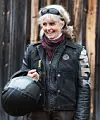 En leende kvinna i skinnjacka och väst håller en svart motorcykelhjälm framför en trävägg.