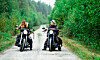 Två personer på motorcyklar sida vid sida på en grusväg kantad av tät grön skog.