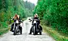 Två personer på motorcyklar sida vid sida på en grusväg kantad av tät grön skog.