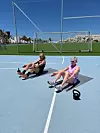 To kvinder laver siddende mavetræning med kettlebells på en udendørs sportsbane.