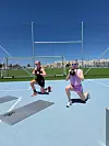 To personer laver lunges med kettlebell på en lys blå udendørs sportsbane i solskin.