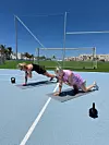 To kvinder træner planke med skulderklap på måtter på en udendørs bane med kettlebells omkring.