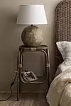Bordslampa på flätat sidobord bredvid en säng med ljusa sängkläder.