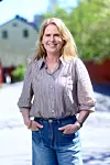 En leende kvinna står utomhus i solsken, klädd i randig skjorta och jeans.