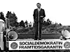 En man står på en upphöjd plattform med högtalare och en skylt med texten ”Socialdemokratisk framtidsgaranti”.