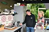 Två personer vid en kaffestation med kaffemaskin, kyl och bord i ett öppet utomhusutrymme.