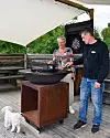 Två personer vid en stor grill på en träaltan under en vit markis, med en liten hund i förgrunden.