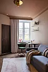 Ljust rum med skrivbord vid fönstret, grå soffa, hyllor på väggen och en matta på golvet.