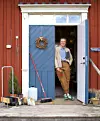 En person står i dörröppningen till en rödmålad byggnad med blå dörrar, kvast och träföremål vid entrén.
