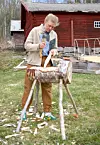 En person täljer trä vid en stock på bockar framför en röd träbyggnad.