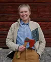 En leende person står framför en röd trävägg och håller en yxa med träskaft.