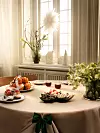 Dukat bord med fruktfat, bakverk, glas och gröna växter framför ett solbelyst fönster.