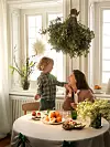 Kvinna och barn vid ett runt bord med frukt, bakverk och blommor i ett soligt rum.