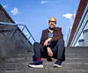 Man i mörk jacka och jeans sitter på en betongtrappa med blå himmel bakom.