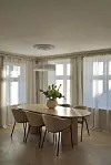 Lyst spisestueinteriør med rundt bord, fire stoler, store vinduer og en vase med blomster.