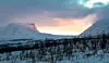 Snötäckta fjäll och mörk skog under en rosa och blå vinterhimmel vid solnedgång.