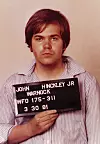 Ung man med kort mörkt hår håller en svart skylt med texten John Hinckley Jr.