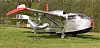 Silverfärgad helikopter med röda vingar står på gräs framför en skog.