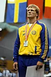 En idrottare i gul blå dräkt står med medalj runt halsen framför svenska flaggor.