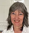 Leende kvinna med grått hår och vit skjorta mot ljus kakelvägg.