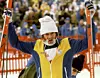 En skidåkare i vit mössa och gulblå jacka håller upp två skidor framför en publikläktare.