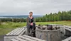 En kvinna sitter på en bänk vid en stenringad grillplats med skog och landskap i bakgrunden.